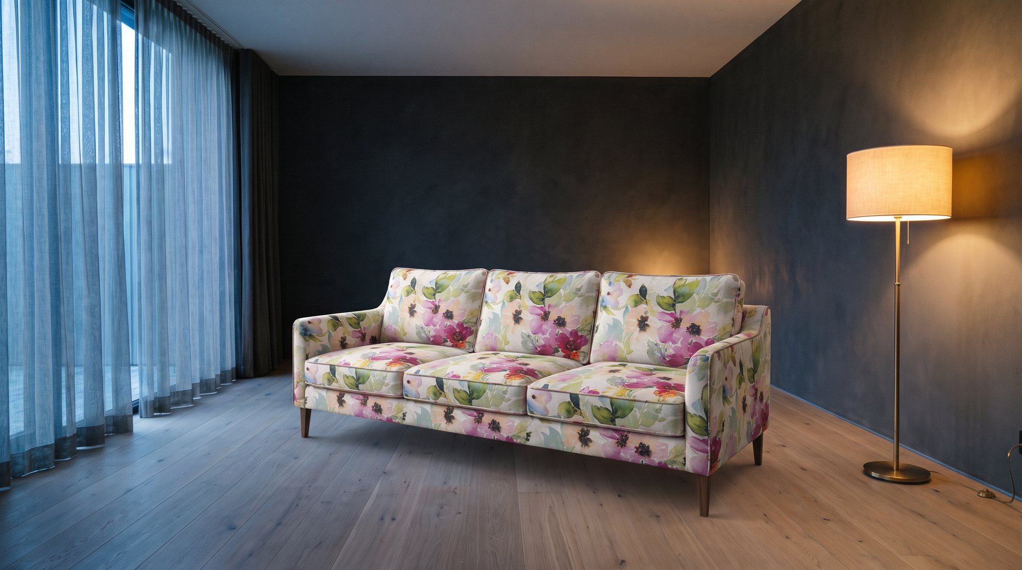 Un sofa haut de gamme retapissé dans un tissu à motif floral aquarelle, dans une pièce sombre éclairée par un rideau voilé et une lampe sur pied.